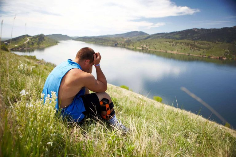 Man sitting in grass on hill beside a lake with head bowed in hands