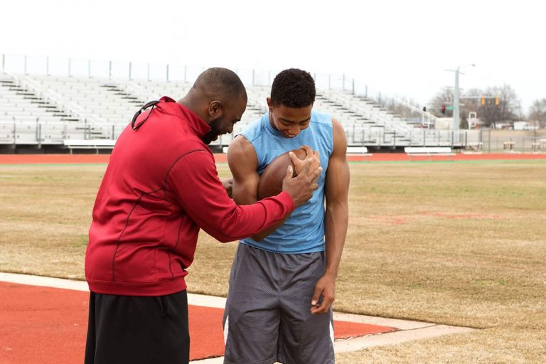 Two men on a track with heads bowed, with one hand each holding a football to the chest of the man in a blue shirt