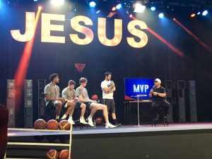 A man is speaking on a stage with three other men seated beside him, with basketballs on the stage and "JESUS" written in large lit letters behind them.