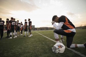 football player on one knee with head bowed at football sideline outside