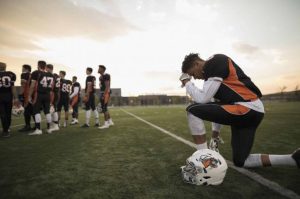 football player on one knee with head bowed at football sideline outside