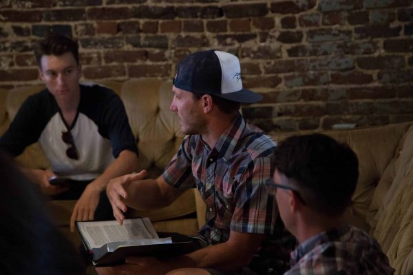 man sitting between two other men and speaking with a Bible in one hand