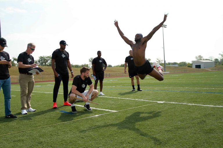 man flying through the air on a grass sports field with several male onlookers.