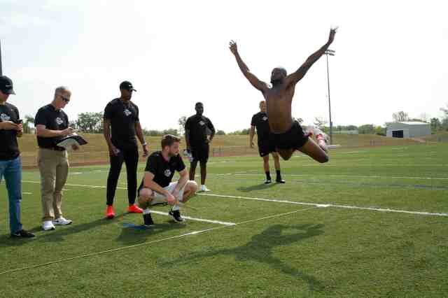 man flying through the air on a grass sports field with several male onlookers.