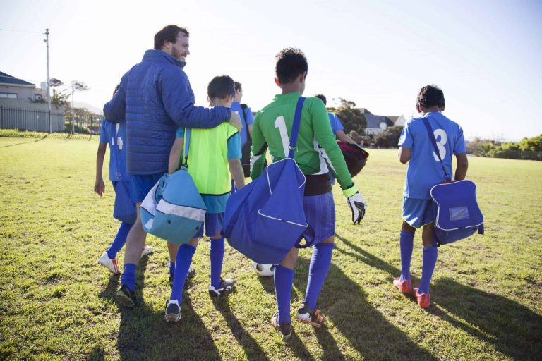 back of group of boys and 1 man with his arm around one of the boys, walking out onto a field carrying their equipment bags