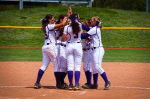 softball team of women in white uniforms huddling in middle of infield giving high fives
