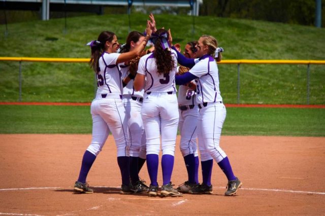 softball team of women in white uniforms huddling in middle of infield giving high fives