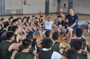 Athletes sitting on basketball court floor looking to a man sitting in a chair speaking.