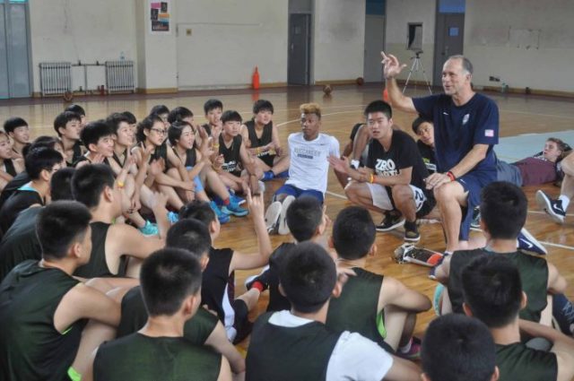 Athletes sitting on basketball court floor looking to a man sitting in a chair speaking.