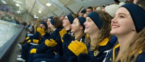 Several women sitting in a row in bleachers all smiling and wearing navy blue headbands, navy-colored uniforms, yellow gloves