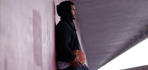 man in black hoodie holding basketball leans against a concrete wall