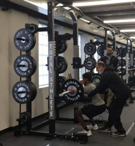 three men in front of weights in weight room, one is squatting