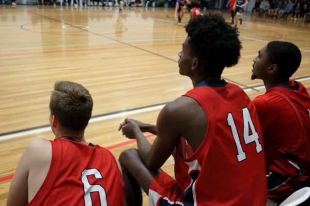 Man in red basketball uniform sitting on sideline of basketball court.