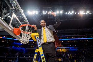 Man with yellow tie standing on a ladder, holding up a piece of net cut from the basketball net next to him