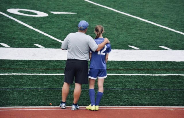 Backs of man with hand on female's shoulder standing on side of playing field.