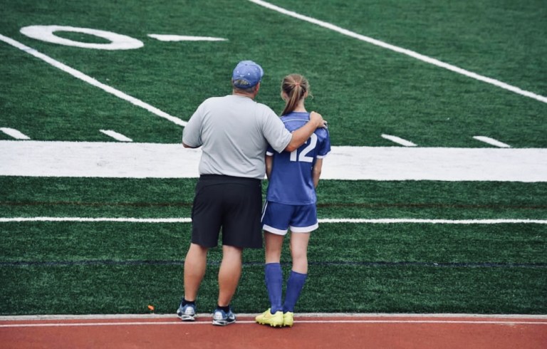 Backs of man with hand on female's shoulder standing on side of playing field.