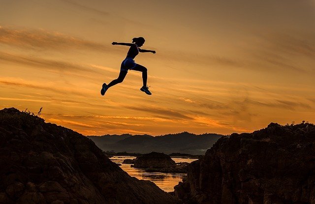 silhouette of a female jumping high in air between two mountains with orange sky and lake in background