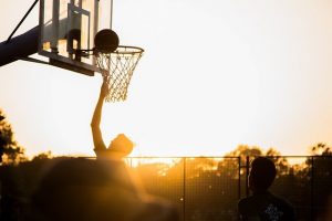 basketball player with hand up and ball above the net on an outdoor court with sun on horizon behind him