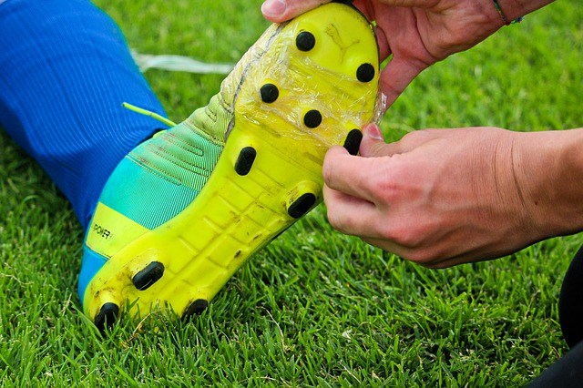 one hand on the top and one hand touching a spike on the bottom of a shoe worn by someone sitting on grass