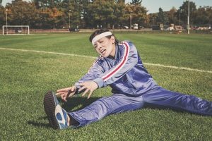 Person with white headband and blue sweats is sitting on grass in field and stretching for a foot.