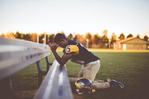 Man kneeling before a sports bench on a field with hands folded prayer-like