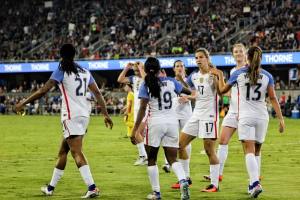 Female athlete in white uniform talking to her team on a field as they are walking together before a large crowd.