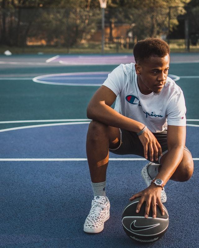 Man squatting down, one elbow on a knee, one hand on a basketball, looking down to the side on an outside court..