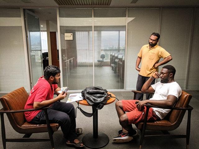 Two men sitting facing each other with a small table between them and one man standing looking on.