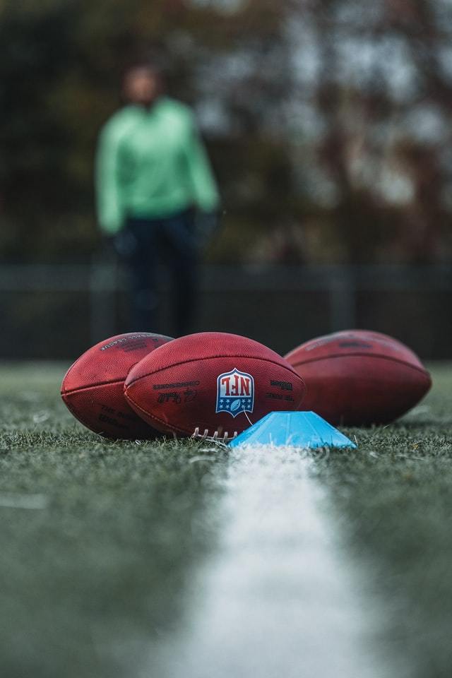 person in aqua jacket looking from sidelines at 3 footballs lying on a yard line in middle of field