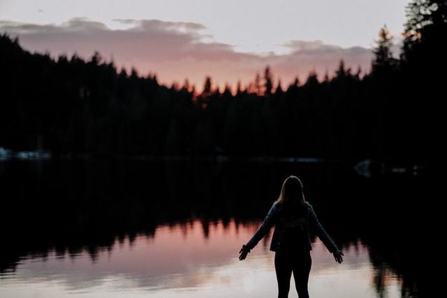 Silhouette from back of a female in shorts with arms out to the side in front of a lake and silhouette of trees and mountains.