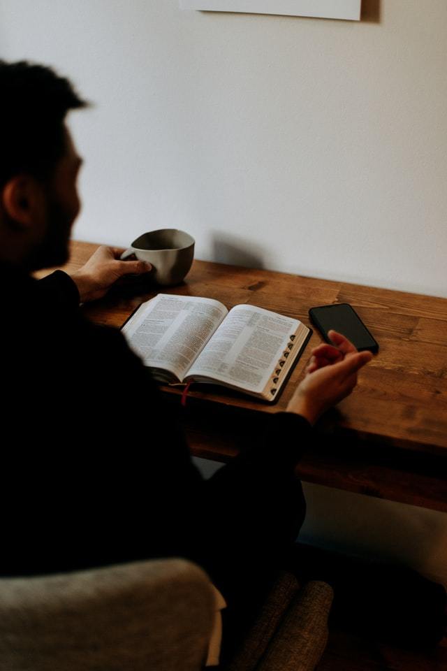 Looking over the shoulder of a shadowy man reading a Bible at a desk.