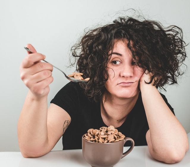 Facing a girl with dark curly hair with elbows on a table, looking at spoonful of food.