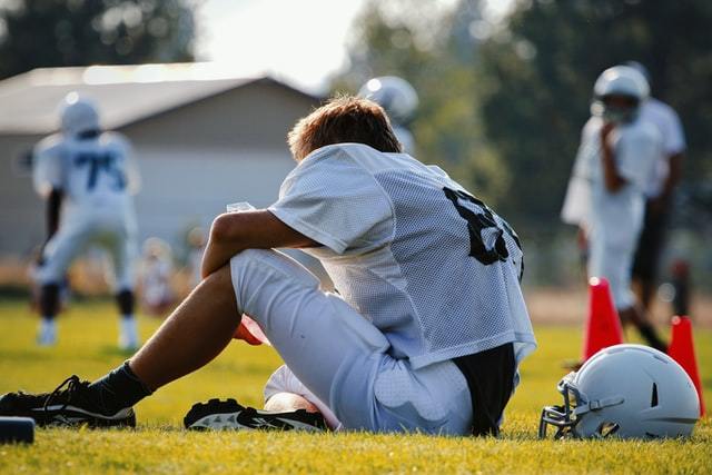 man sitting on sideline of football game in a uniform, helmet laying next to him