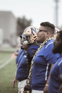 men, some in football gear, standing on sidelines