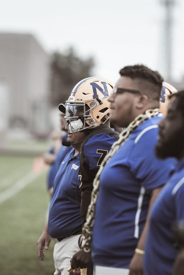 men, some in football gear, standing on sidelines