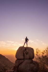 Man with his back to the camera looking toward the horizon while standing on top of a large rock.