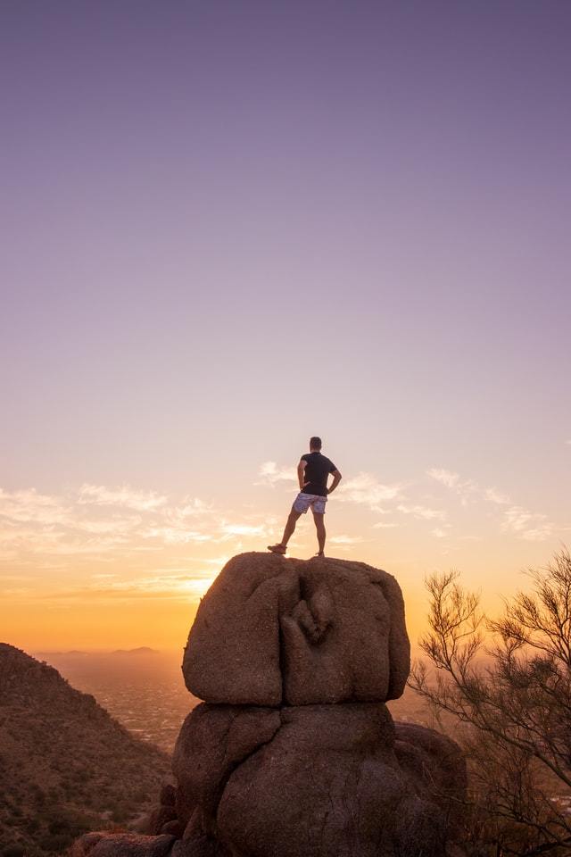 Man with his back to the camera looking toward the horizon while standing on top of a large rock.