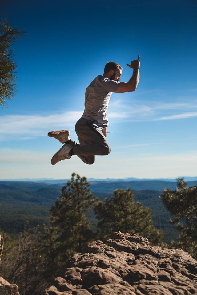 Man jumping high above rocks, trees, mountains and skyline.