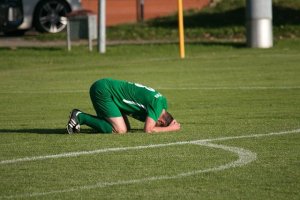 athlete in green uniform kneeling on field with his head down and hands on head