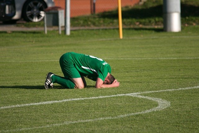 athlete in green uniform kneeling on field with his head down and hands on head