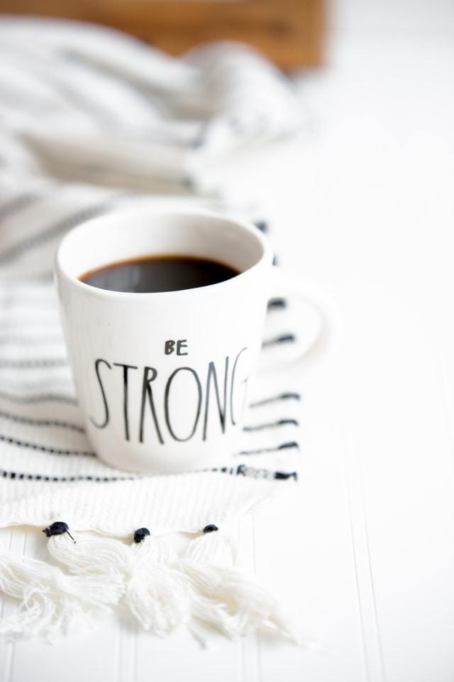 white mug that says "BE STRONG" with a dark liquid in it .
