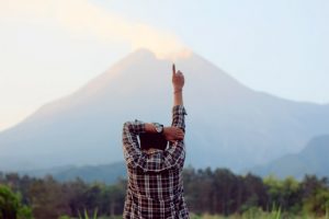 Person standing in front of a volcano with 1 arm bent over head and 1 arm and finger pointing up