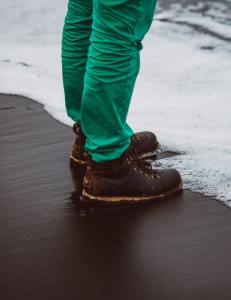 legs and feet from knee down in green pants, brown boots, standing on cement partly covered in snow.
