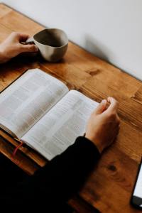 hand holding a cup, other hand touching a Bible while sitting at a table