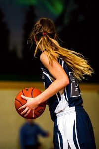 girl with long hair in a ponytail on a basketball court holding a basketball, looking downcourt away from camera