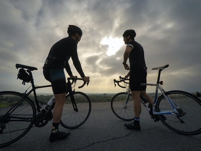 Two riders stopped on bicycles facing each other with clouds and sun rays behind them
