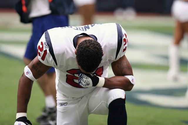 Facing a football player kneeling on one knee on field with head bowed, one arm resting on raised knee.