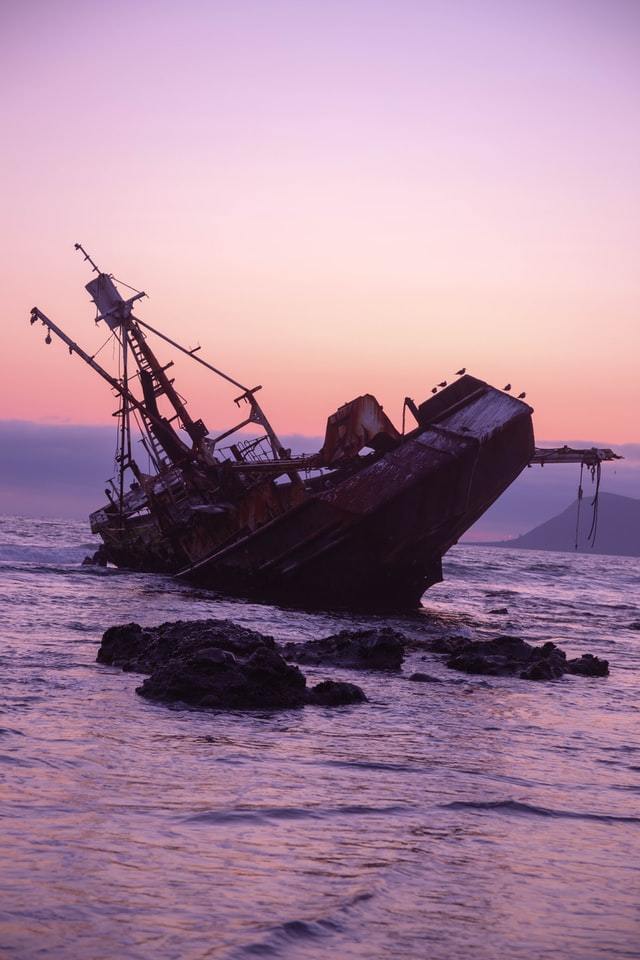 Ship listing to the left, half sunk in the water with pinkish sky behind it.