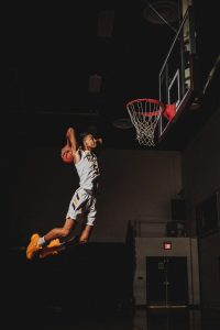 Basketball player in white uniform is flying through the air in front of a basketball net, with a basketball cocked behind his head ready to dunk it.