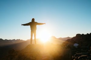 Man standing on a hill with arms outstretched gazing toward the sun on the horizon.
